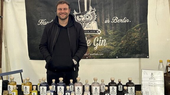 Der Gründer von Gentle Gin, Stanislav Bic, steht in schwarzer Bekleidung an einem Marktstand. Mehrere Flaschen Gin sind vor ihm aufgereiht.