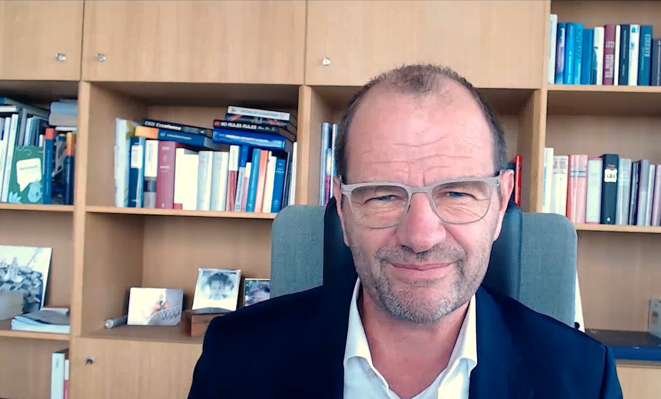 Zu sehen ist ein mittelalter Mann mit Brille, der vor einem Bücherregal sitzt und einen Anzug trägt.