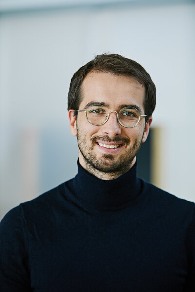 Ein Mann mit runder Brille und schwarzem Rollkragenpullover lächelt in die Kamera. Im Hintergrund ist eine unscharfe Kulisse zu sehen.