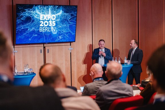 Zwei Männer unterhalten sich vor Publikum auf einer Bühne.