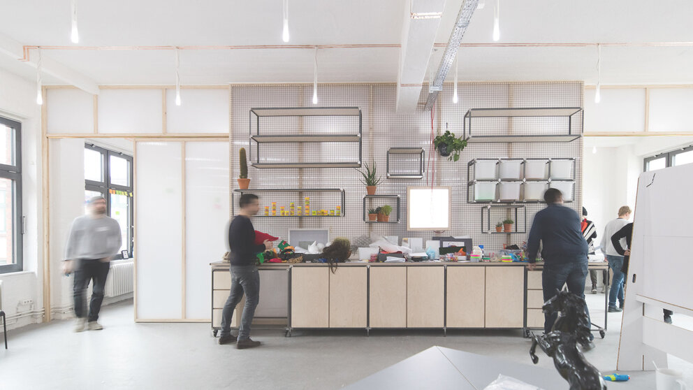 Innenraum eines hellen, modernen Workspaces, in dem mehrere Menschen stehend arbeiten. 