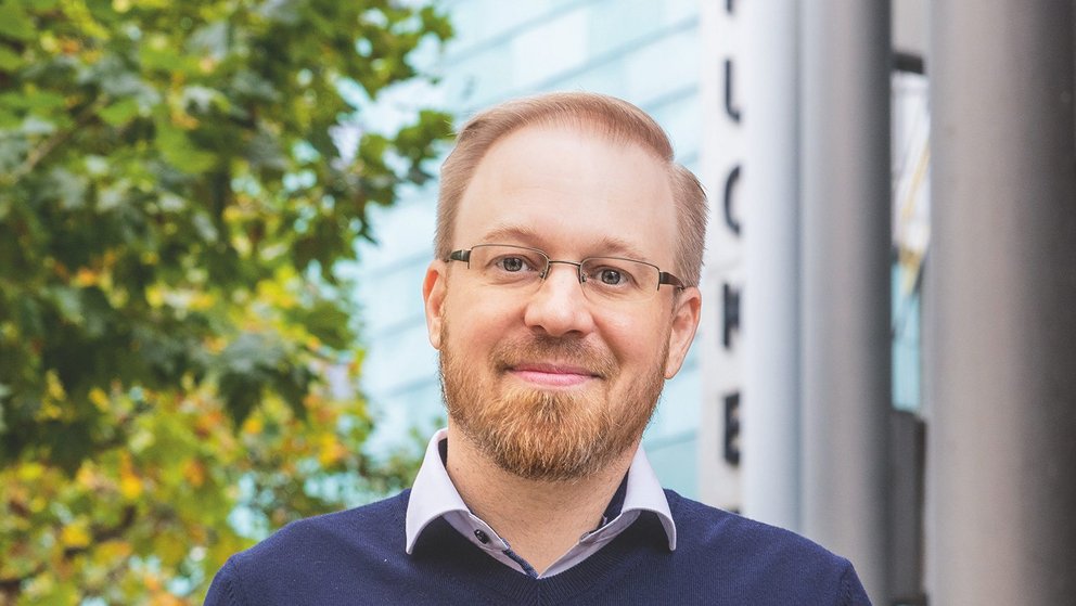 Das Bild zeigt einen rotblonden Mann mit Bart und Brille mit blauem Pullover und hellem Hemd