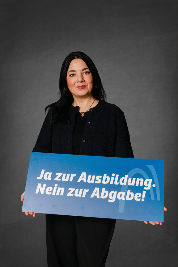 Eine Person steht vor einem grauen Hintergrund und hält ein blaues Schild mit weißer Schrift.