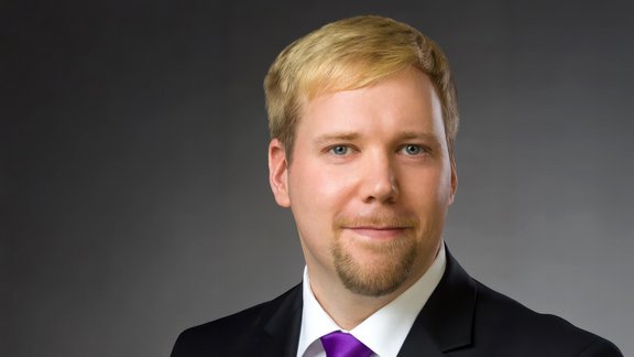 Mann mit kurzem blondem Haar trägt schwarzen Anzug, weißes Hemd und schwarze Krawatte vor grauem Hintergrund.