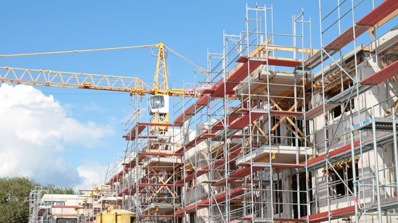 Zu sehen ist der Rohbau eines Geschosshauses. Um das Gebäude herum ist ein Baugerüst angebracht, im Hintergrund steht ein großer gelber Baukran.