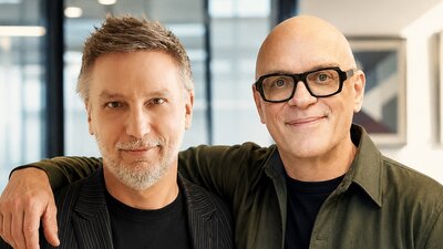 Auf dem Bild sin zwei Männer zu sehen. Beide stehen relativ nah an der Kamera. Der Mann rechts hat seinen Arm um den Mann links. Beide lächeln in die Kamera