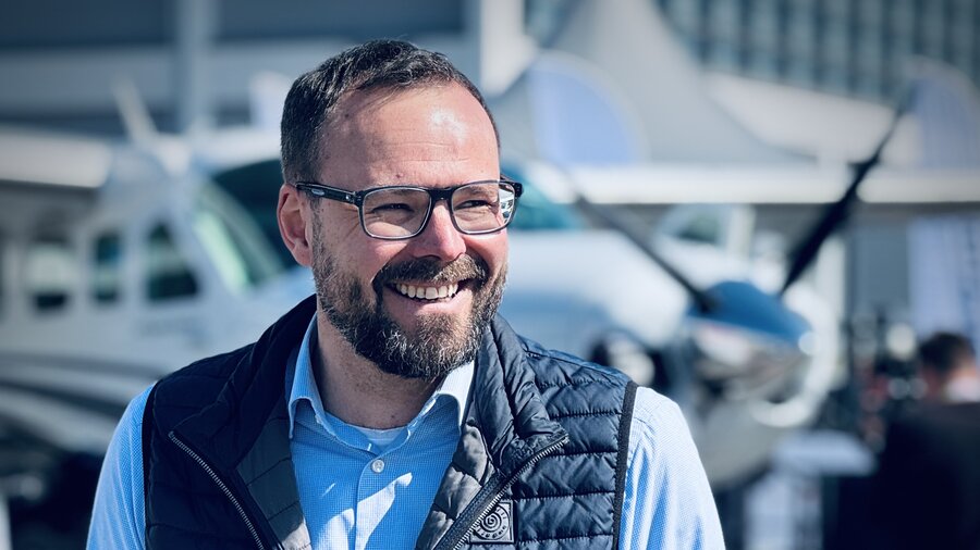 Zu sehen ist ein Mann mit Brille und Bart vor dem Hintergrund eines Kleinflugzeugs, es ist Fabian Rosenberg, Geschäftsführer der AIR UNIQON GmbH
