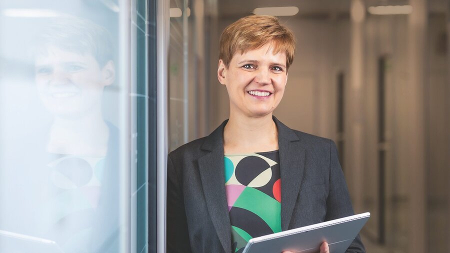 Zu sehen ist eine dunkelblonde Frau mit dunkler Jacke und farbiger Bluse.