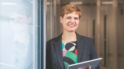 Zu sehen ist eine dunkelblonde Frau mit dunkler Jacke und farbiger Bluse.
