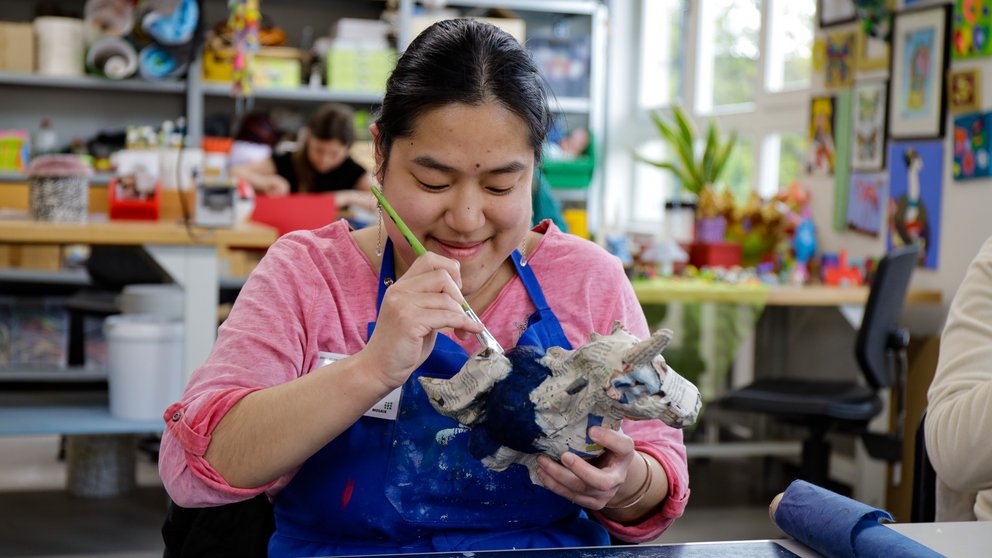 Person mit rosa Hemd und blauer Schürze bemalt eine weiße, mehrköpfige Drachenfigur aus Ton in einem hellen, mit Kunstwerken dekorierten Atelier.