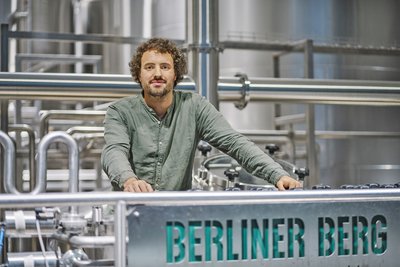 Person mit lockigem Haar und grünem Hemd steht an einer Brauereianlage mit der Aufschrift 'BERLINER BERG'
