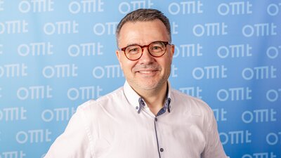 Ein Mann mit Brille steht vor einer hellblauen Leinwand. Er trägt ein weißes Hemd.