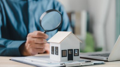 Zu sehen ist ein Schreibtisch auf dem ein Laptop steht und ein Klemmbrett liegt. Auf dem Klemmbrett steht ein Modellhaus. Eine Person richtet eine Lupe auf das Modellhaus.