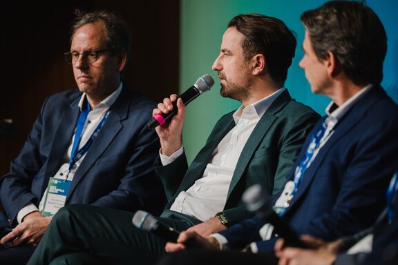 Robert Rückel spricht auf einer Bühne sitzend in ein Mikrofon.