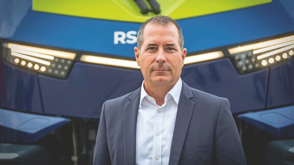 Ein Mann mittleren Alters steht mit blauem Jaket und hellem Hemd vor einem Fahrzeug. 