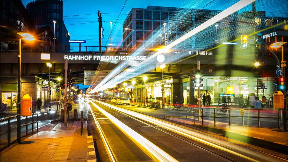 Zu sehen ist eine Straße sowie langgezogene Lichter durch eine Langzeitbelichtung. Es handelt sich um die Friedrichstraße in Berlin.