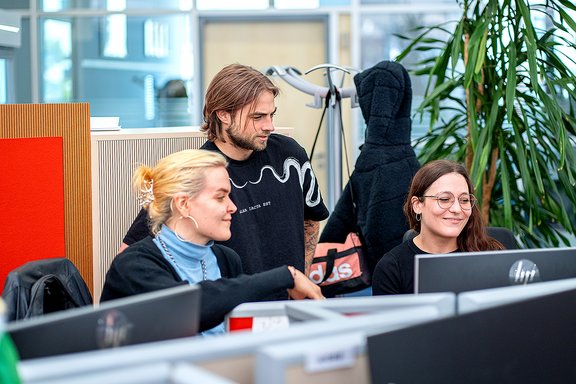 Drei junge Erwachsene arbeiten gemeinsam an Computern in einem modernen Büro mit Pflanzen und Garderobe im Hintergrund