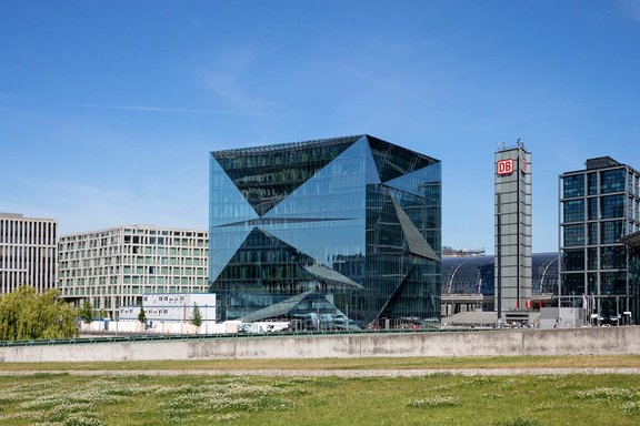 Das Cube Berlin, ein würfelförmiges Glasgebäude, umgeben von weiteren modernen Bürogebäuden am Spreeufer