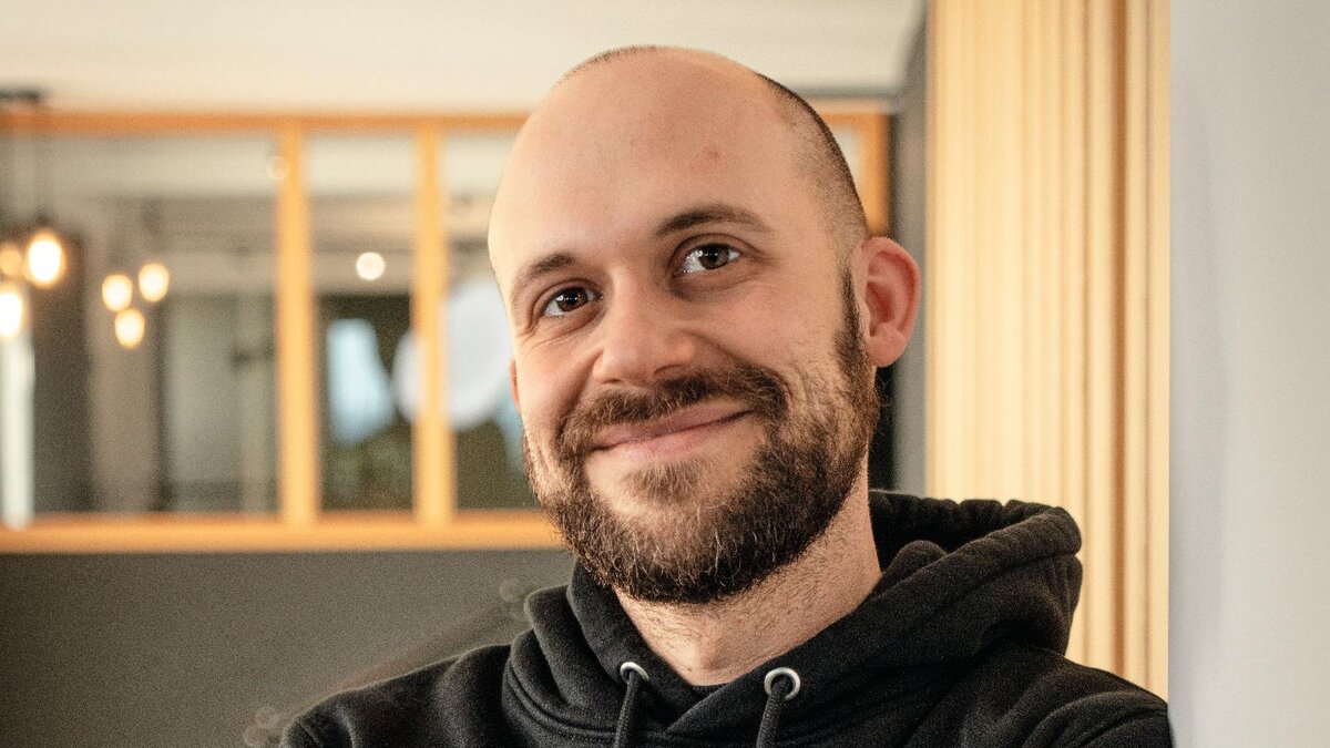 Zu sehen ist ein junger Mann mit Glatze, welcher einen schwarzen Kapuzenpollover trägt. Er verschränkt die Arme vor seiner Brust und schaut freundlich in die Kamera.