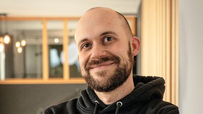 Zu sehen ist ein junger Mann mit Glatze, welcher einen schwarzen Kapuzenpollover trägt. Er verschränkt die Arme vor seiner Brust und schaut freundlich in die Kamera.