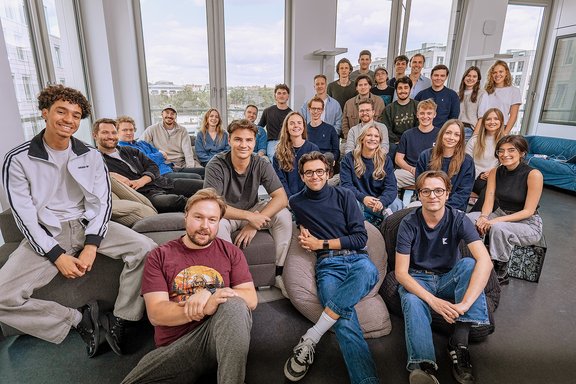 Gruppe von jungen Erwachsenen sitzt und steht in einem modernen Raum mit großen Fenstern und Blick auf eine Stadtlandschaft.
