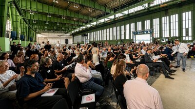 Menschen sitzen in Stuhlreihen in einer früheren Industriehalle, es ist die Eröffnung der Ausstellung „Ring frei für eine IBA!“