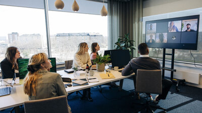 Kollegen sitzen in einem Meetingraum – weitere sind digital zugeschaltet.