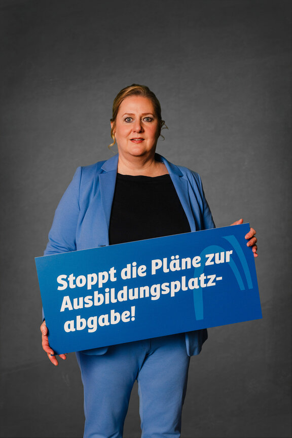 Eine Person steht vor einem neutralen grauen Hintergrund und hält ein großes blaues Schild mit der weißen Aufschrift: "Stoppt die Pläne zur Ausbildungsplatzabgabe!"