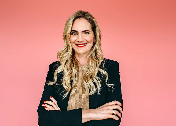 Frau mit langen, welligen blonden Haaren, gekleidet in einem schwarzen Blazer und einem beigen Pullover, steht mit verschränkten Armen vor einem rosa Hintergrund.