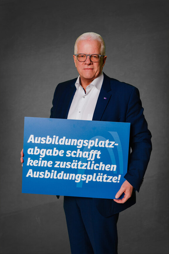 Eine Person steht vor einem neutralen grauen Hintergrund und hält ein großes blaues Schild mit der weißen Aufschrift: "Ausbildungsplatzabgabe schafft keine zusätzlichen Ausbildungsplätze!"