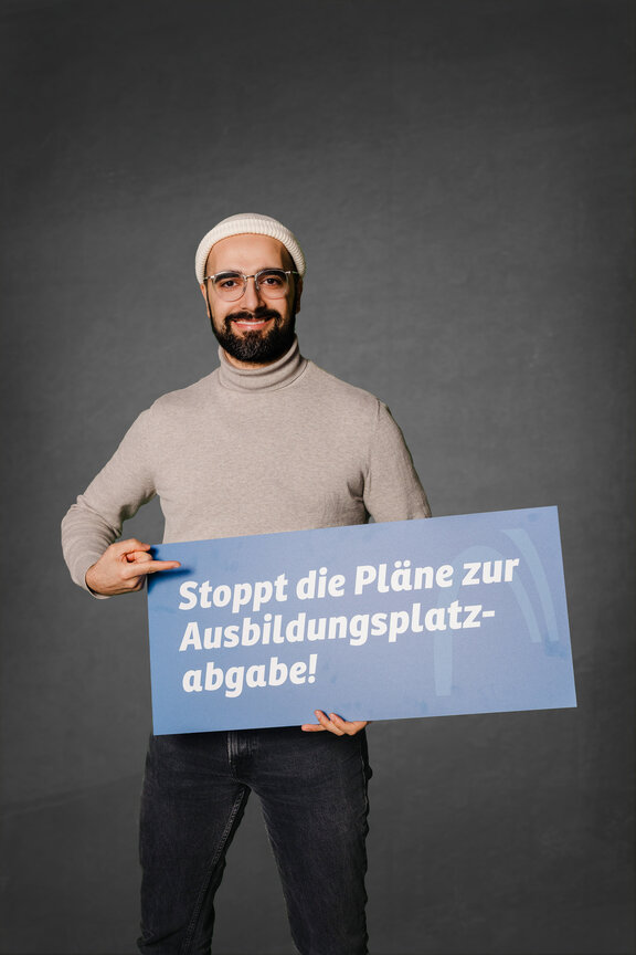 Eine Person steht vor einem neutralen grauen Hintergrund und hält ein großes blaues Schild mit der weißen Aufschrift: "Ja zur Ausbildung. Nein zur Abgabe!"