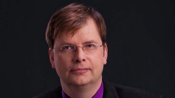 Ein mittelalter Mann mit Brille und lila Hemd vor einem schwarzen Hintergrund. Es ist der Generaldirektor des Robotik-Unternehmens pi4.