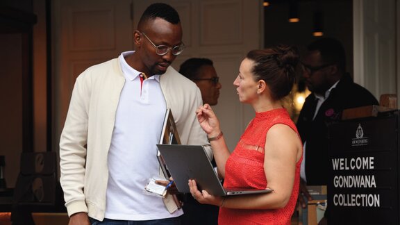 Zu sehen ist eine Frau in einem roten Kleid. Sie hält einen aufgeklappten Laptop und unterhält sich mit einem Mann, der neben ihr steht und ein weißes Shirt sowie eine weiße Jacke trägt.