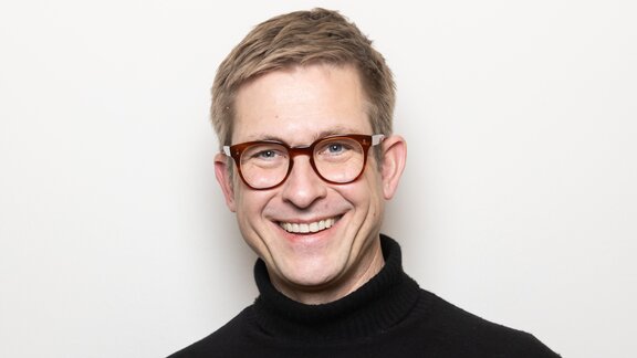 Ein Mann mit kurzen Haaren und Brille lächelt freundlich in die Kamera 