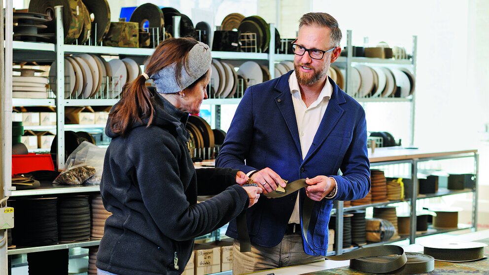 Zwei Menschen im Gespräch vor einer Werkbank. Beide halten einen Stoff in der Hand 