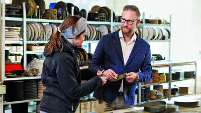 Zwei Menschen im Gespräch vor einer Werkbank. Beide halten einen Stoff in der Hand 