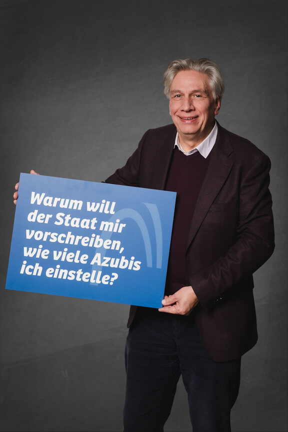 Eine Person steht vor einem neutralen grauen Hintergrund und hält ein großes blaues Schild mit der weißen Aufschrift: "Warum will der Staat mir vorschreiben, wie viele Azubis ich einstelle?""