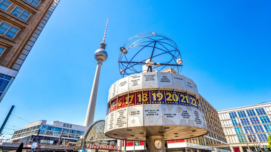 Zu sehen ist ein Turm, Gebäude und eine Uhr. Es handelt sich um den Alexanderplatz in Berlin mit der Weltzeituhr und dem Fernsehturm.