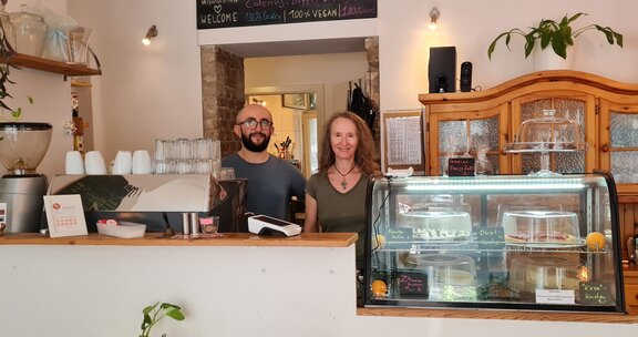 Eine Frau und ein Mann stehen hinter einem weißen Tressen in einem Cafe. Zu sehen sind mehrere helle Holzmöbel und grüne Pflanzen 