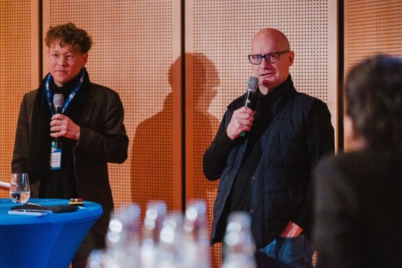 Zwei Männer halten ein Mikrofon, einer spricht.