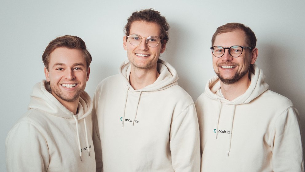 Drei Männer in beigen gleichfarbigen Sweatpullovern stehen nebeneinander und lächeln in die Kamera. 