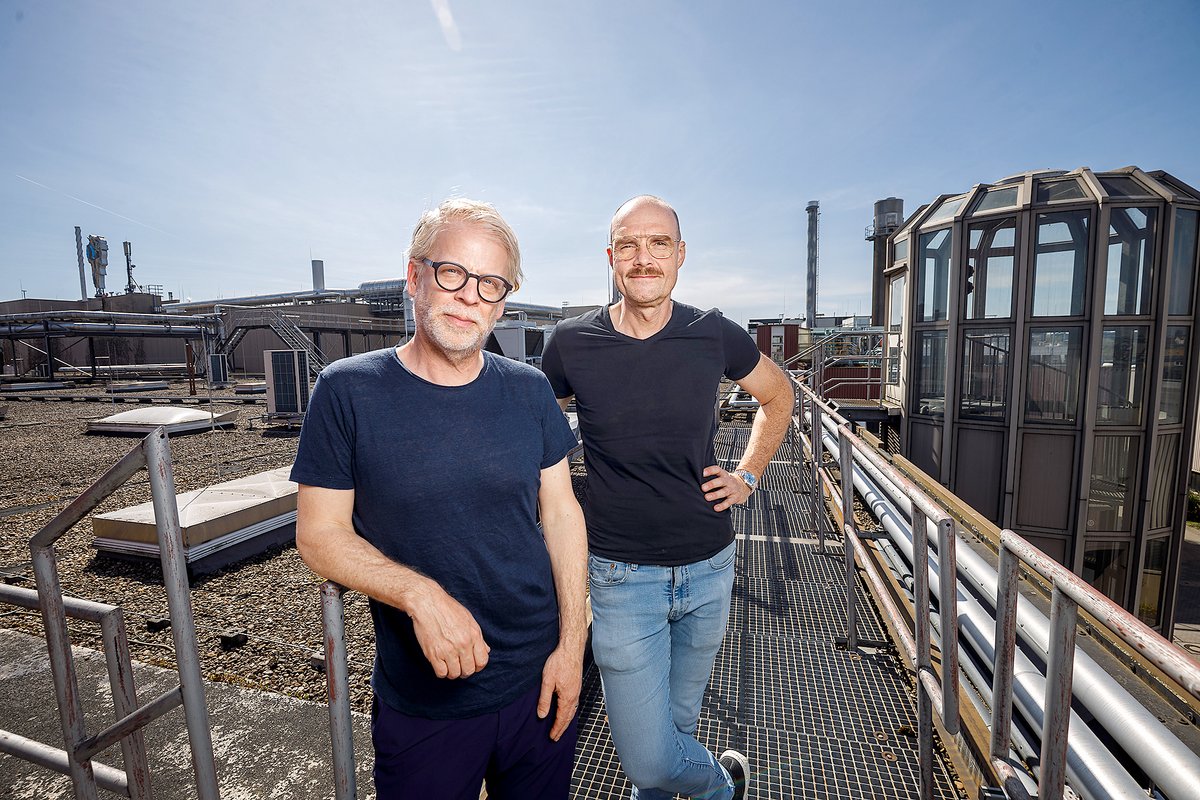 Zwei Männer mit Brillen und schwarzen T-Shirts stehen auf einem Dach mit Industrieanlagen im Hintergrund, sonniger Himmel