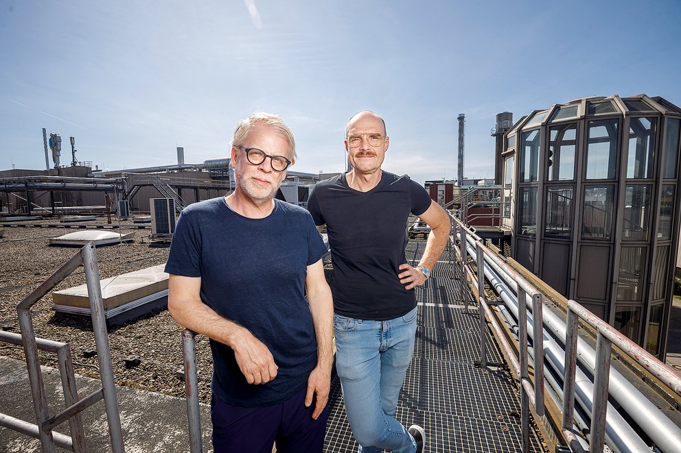 Zwei Männer mit Brillen und schwarzen T-Shirts stehen auf einem Dach mit Industrieanlagen im Hintergrund, sonniger Himmel
