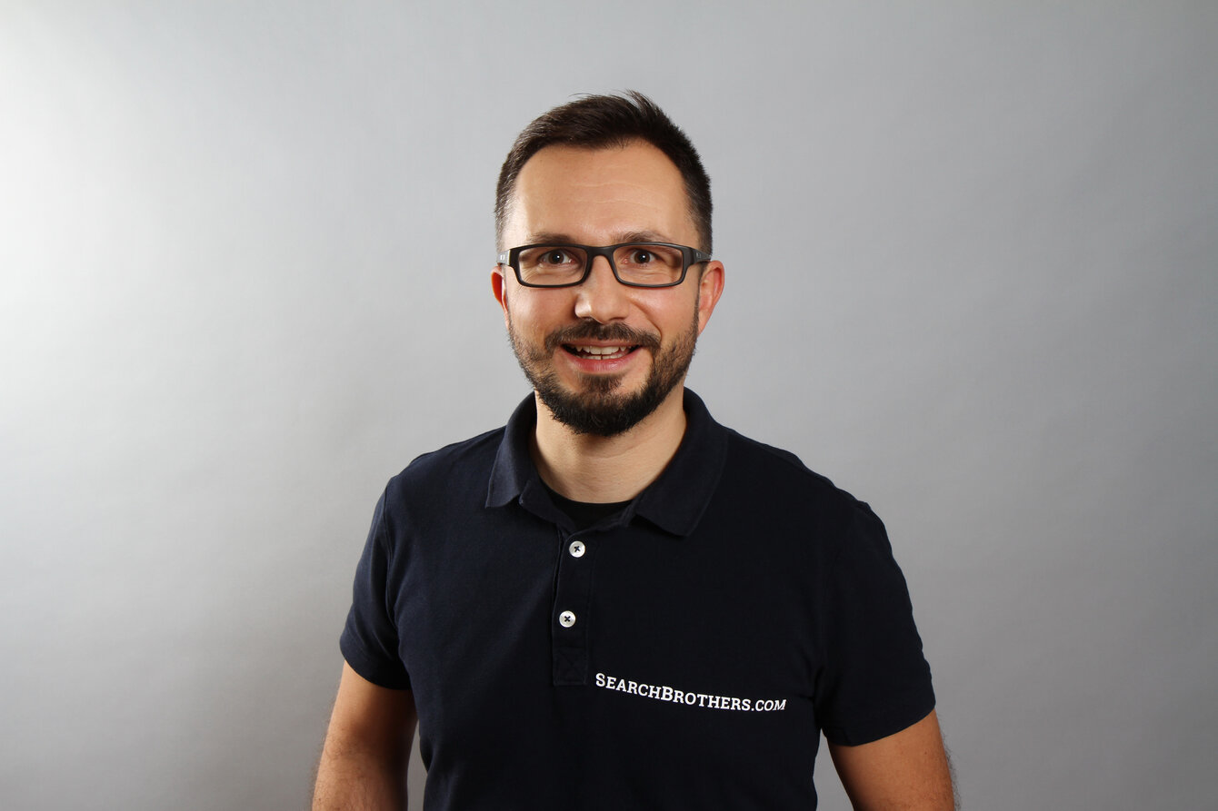 Professionelles Porträt einer Person in dunklem Poloshirt vor neutralem grauem Hintergrund. Zu sehen ist Kaspar Szymanski von Search Brothers