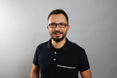 Professionelles Porträt einer Person in dunklem Poloshirt vor neutralem grauem Hintergrund. Zu sehen ist Kaspar Szymanski von Search Brothers