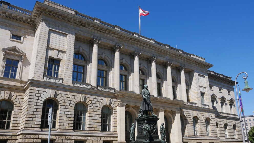 Zu sehen ist ein großes, helles, imposantes Gebäude. Es ist sehr breit, zweistöckig und auf dem Dach weht die Fahne Berlins. Es handelt sich um das Abgeordnetenhaus Berlins. Vor dem Haus steht eine Statue.