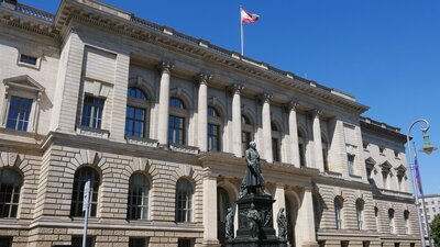 Zu sehen ist ein großes, helles, imposantes Gebäude. Es ist sehr breit, zweistöckig und auf dem Dach weht die Fahne Berlins. Es handelt sich um das Abgeordnetenhaus Berlins. Vor dem Haus steht eine Statue.