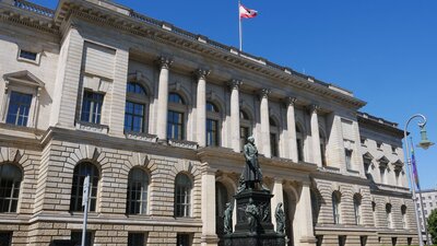 Zu sehen ist ein großes, helles, imposantes Gebäude. Es ist sehr breit, zweistöckig und auf dem Dach weht die Fahne Berlins. Es handelt sich um das Abgeordnetenhaus Berlins. Vor dem Haus steht eine Statue.