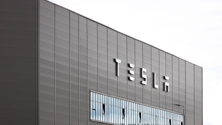 Zu sehen ist ein graues blockartiges Gebäude mit der Aufschrift Tesla. Es handelt sich um die Gigafactory des Autoherstellers in Grünheide, Brandenburg.