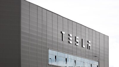 Zu sehen ist ein graues blockartiges Gebäude mit der Aufschrift Tesla. Es handelt sich um die Gigafactory des Autoherstellers in Grünheide, Brandenburg.